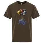 T-shirt marron avec un motif d'astronaute flottant portant un ballon formé de plusieurs planètes colorées, créant une scène spatiale ludique et imaginative.