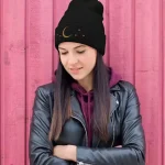 Jeune femme portant un bonnet noir avec motif lunaire et étoiles, vêtue d'une veste en cuir noire et d'un sweat à capuche violet, posant devant un mur en bois rose.