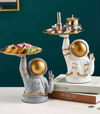 Figurine d'astronaute en céramique blanche et dorée, utilisée comme décoration, tenant un plateau avec des objets de décoration et des bonbons, style moderne et original.