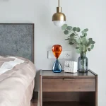 Vase en verre orange et bleu sur une table de nuit moderne avec plante verte et lampe suspendue dorée.