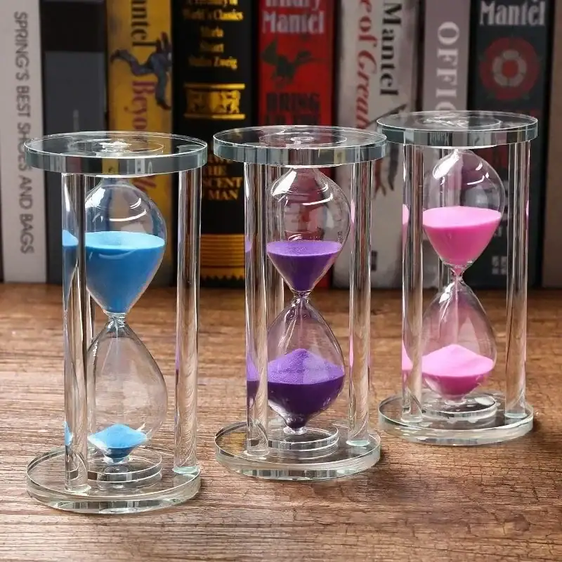 sablier-en-verre-de-cristal-1 Sablier coloré en verre avec trois tubes contenant du sable bleu, violet et rose, placé sur une surface en bois avec des livres en arrière-plan.