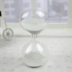Sablier en verre avec sable blanc, symbole du temps qui passe, posé sur une surface claire avec un décor minimaliste en arrière-plan.