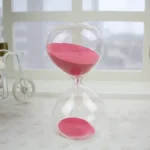 Sablier en verre transparent avec sable rose, symbole du temps qui passe, posé sur une surface claire avec un décor minimaliste en arrière-plan.