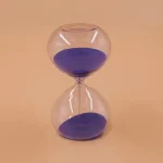 Sablier en verre transparent avec sable violet, symbole du temps qui passe, posé sur un fond beige.