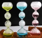 Balançoire en verre coloré avec bulles d'air, composée de trois segments en forme de boule, chacun rempli de liquide coloré (jaune, bleu, rose) et de bulles d'air, sur fond noir.