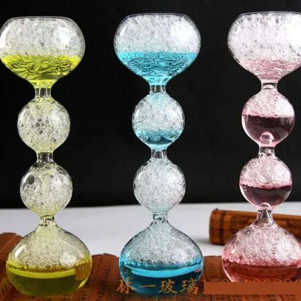 Balançoire en verre coloré avec bulles d'air, composée de trois segments en forme de boule, chacun rempli de liquide coloré (jaune, bleu, rose) et de bulles d'air, sur fond noir.
