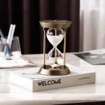 Sablier de décoration en laiton et verre, placé sur un livre intitulé "WELCOME" dans un environnement de bureau ou de salon.