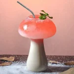Verre en forme de champignon contenant un cocktail rose avec une branche de menthe, décoratif et original.