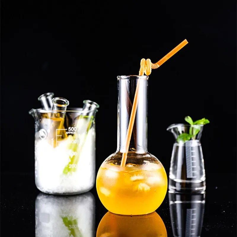 verre-a-cocktail-design-de-chimiste-1 Vase de laboratoire contenant une boisson orange avec une paille, accompagné d'autres accessoires de laboratoire en verre, sur fond noir.