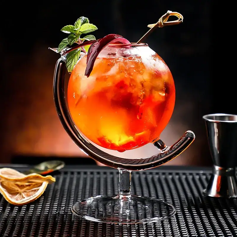 verre-a-cocktail-globe-terrestre-1 Cocktail coloré dans un verre original avec garniture de menthe et tranche de citron, présenté sur un tapis noir avec accessoires de bar en arrière-plan.