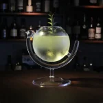 Cocktail lumineux dans un bocal scientifique avec une plante verte flottante, présenté sur un support en verre, ambiance de bar avec étagères de bouteilles en arrière-plan.