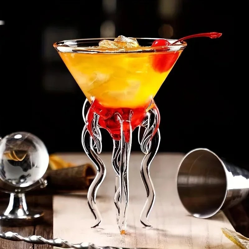 verre-a-cocktail-meduse-1 Verre à cocktail scientifique avec décor en forme de tentacules en verre transparent, rempli d'un cocktail jaune-orange avec cerise, ambiance de bar moderne.