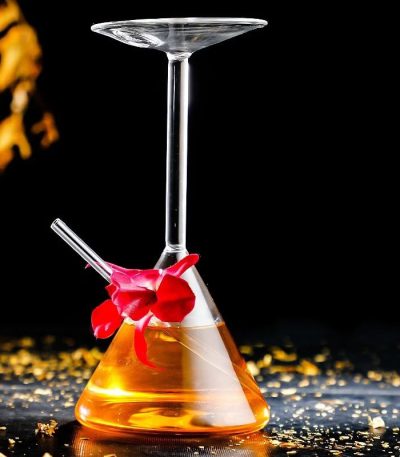 Béchier de laboratoire avec verre à cocktail suspendu, contenant un liquide orange, décoré d'une fleur rouge, sur une surface noire avec des éclats dorés.
