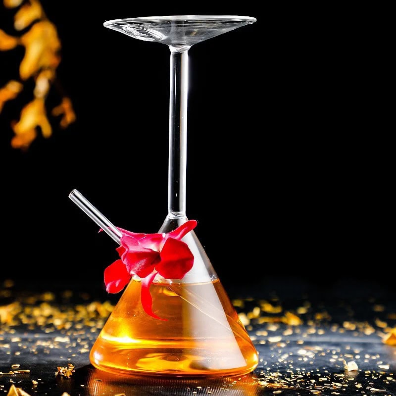 verre-a-cocktail-original-erlenmeyer-1 Béchier de laboratoire avec verre à cocktail suspendu, contenant un liquide orange, décoré d'une fleur rouge, sur une surface noire avec des éclats dorés.