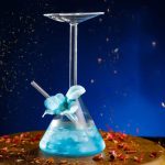 Verrerie scientifique contenant un liquide bleu lumineux, avec un ballon à décanter et un tube en verre, sur un fond sombre avec des éclats de lumière et des pétales.