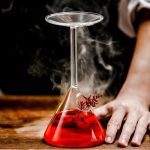 Flacon de laboratoire en verre contenant un liquide rouge fumant, posé sur une surface en bois, avec une main à côté, illustrant une expérience scientifique.