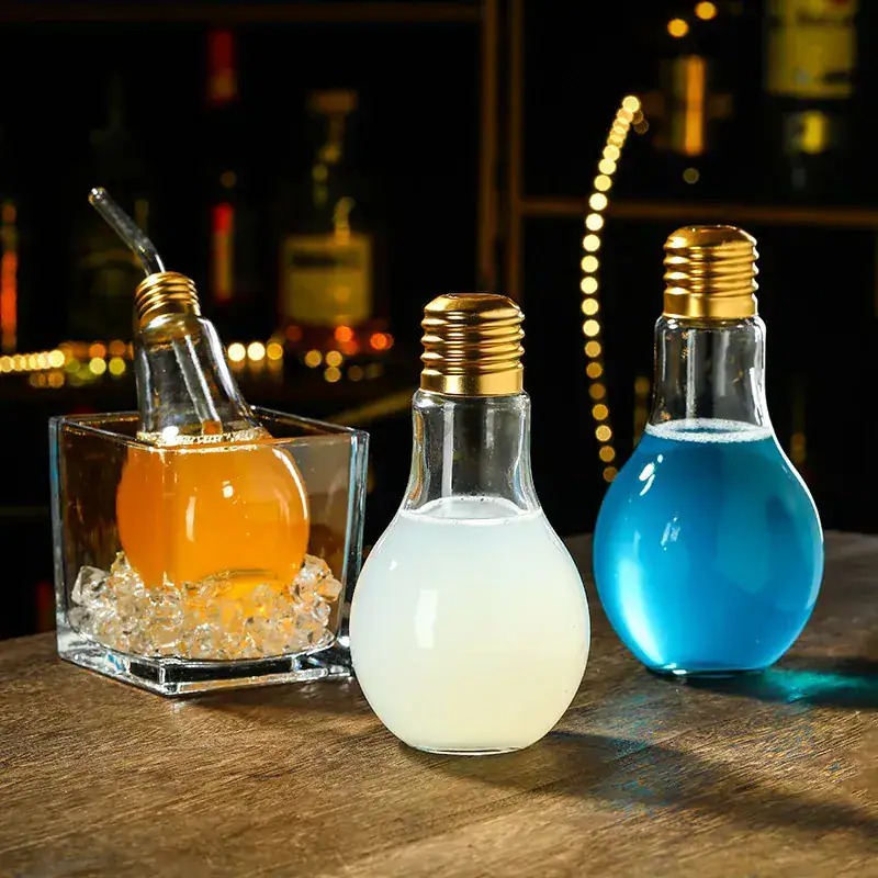 verre-ampoule-cocktail-1 Trois ampoules décoratives remplies de liquides colorés (orange, blanc, bleu) sur une table en bois, ambiance chaleureuse et moderne.