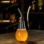 Ampoule décorative en forme de bouteille avec boisson orange, équipée d'une paille métallique, sur une surface en bois dans un environnement sombre et moderne.