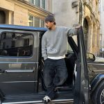 Homme en tenue décontractée posant devant une voiture noire dans une rue urbaine.